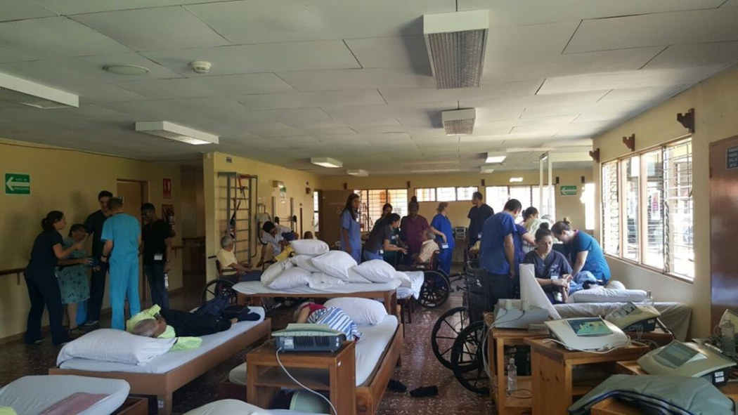 Busy hospital ward with patients in beds, nurses and volunteers assisting along the aisle amid wheelchairs and medical equipment.